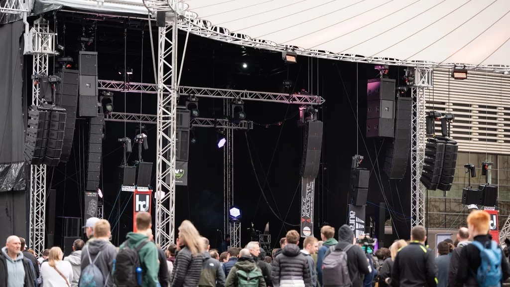 The image shows a large outdoor stage with a professional PA sound system at the Prolight + Sound trade fair. The stage is surrounded by a sturdy truss system from which several line array speaker systems from various manufacturers are suspended. In the foreground is a crowd listening to the presentations or waiting for the next demonstrations. The atmosphere feels dynamic and demonstrates how the Live Sound Arena serves as a central platform for audio technology enthusiasts and trade visitors. The logos of participating manufacturers such as ‘hd’ (Harmonic Design) and ‘Voice-Acoustic’ are visible on the loudspeaker systems, indicating that the latest sound reinforcement technologies are being showcased here.