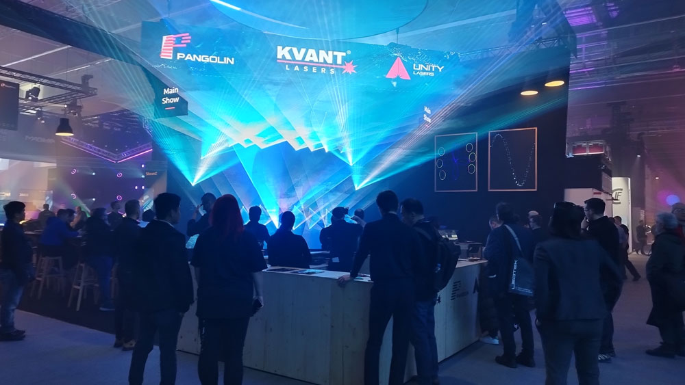 At Prolight + Sound 2024, a large crowd of visitors gather around the KVANT stand, where impressive blue laser beams form an impressive triangular light display above the area. The logos of KVANT, Pangolin and Unity Lasers are displayed above the installation. The scene exudes a vibrant trade fair atmosphere, with a knowledgeable audience attentively following the laser presentation.
