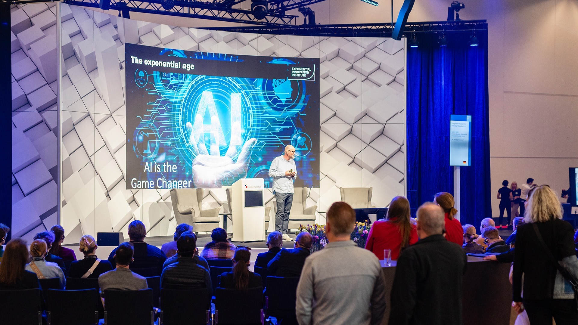 A speaker presents on a stage on the topic of AI in front of an attentive audience.