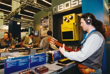 Das Bild zeigt eine Szene von einem Messestand, der dem Hersteller "Boss" gewidmet ist. Im Vordergrund ist ein Mann mit langen Haaren zu sehen, der Kopfhörer trägt und auf einer akustischen Gitarre spielt. Der Fokus liegt auf der Interaktion mit den präsentierten Produkten, darunter Effektgeräte wie der "Super OverDrive SD-1", der in übergroßer Darstellung an der Wand hängt. Im Hintergrund spielen weitere Personen elektrische Gitarren, ebenfalls mit Kopfhörern ausgestattet, um die vorgestellten Audio- und Effektgeräte auszuprobieren. Auf dem Tisch befinden sich mehrere Produkte und Beschreibungen, die die Funktionsweise und Eigenschaften der Geräte erläutern. Die Szenerie vermittelt eine aktive, praxisorientierte Atmosphäre, in der Besucher die Gelegenheit haben, Equipment in einer professionellen Umgebung zu testen.