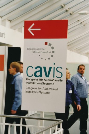 Das Bild zeigt ein Hinweisschild innerhalb eines modernen Gebäudes mit Glasdach. Das Schild weist auf den "CAVIS – Congress für AudioVisuelle InstallationsSysteme" hin, der im Congress Center der Messe Frankfurt stattfindet. Der Name des Kongresses ist in großen, auffälligen Buchstaben dargestellt, ergänzt durch die englische Übersetzung "Congress for AudioVisual InstallationSystems". Ein roter Pfeil auf dem Schild gibt die Richtung an. Im Hintergrund sind mehrere Personen zu sehen, die in einer geschäftigen Atmosphäre vorbeigehen.