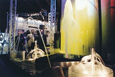 Das Bild zeigt eine kreative Wasserinstallation, die mit bunten Lichtprojektionen kombiniert ist. Im Vordergrund fließt Wasser aus futuristisch gestalteten Brunnen, während sich im Hintergrund eine beleuchtete Wasserwand erstreckt, die in gelblichen und grünen Tönen erstrahlt. Darüber wölben sich elegante Wasserbögen, die harmonisch in das Design eingebettet sind. Im Hintergrund stehen mehrere Personen in Anzügen, die die Installation mit Interesse betrachten. Die Szenerie wird von technischen Bühnenkonstruktionen und weiteren Ausstellungsbereichen umrahmt.