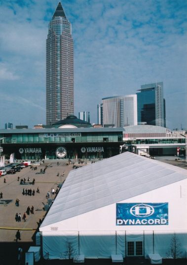 Das Bild zeigt das Messegelände in Frankfurt mit dem markanten Messeturm im Hintergrund, einem der höchsten Wolkenkratzer Deutschlands. Im Vordergrund ist ein großes weißes Zelt mit der Aufschrift „DYNACORD“ sichtbar. Dahinter befindet sich der Eingang des Forums, auf dem große Yamaha-Logos und ein Schriftzug „music4kids“ angebracht sind. Besucher bewegen sich auf dem weitläufigen Platz zwischen den Gebäuden, was die lebhafte Atmosphäre eines Messetages unterstreicht. Der blaue Himmel und die modernen Gebäude geben der Szene einen professionellen und einladenden Charakter.