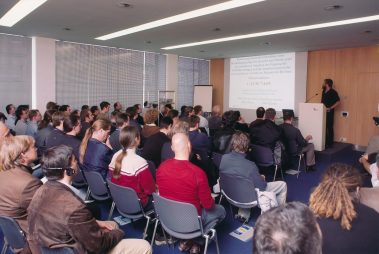Das Bild zeigt einen voll besetzten Konferenzraum, in dem ein Vortrag stattfindet. Die Zuhörer, die unterschiedlich gekleidet sind, hören aufmerksam einem Redner zu, der an einem Pult steht. Auf der Leinwand im Hintergrund ist eine Präsentation mit dem Titel und weiteren Informationen zu einem akustischen Thema, insbesondere zur Beschallung im Berliner Olympiastadion, zu sehen. Die moderne Raumgestaltung mit großen Fenstern und schlichten Möbeln deutet auf ein professionelles Umfeld hin. Der Vortrag scheint Teil einer Fachveranstaltung oder eines Workshops zu sein, wobei das Publikum aus Experten oder Interessierten an der Thematik zu bestehen scheint.
