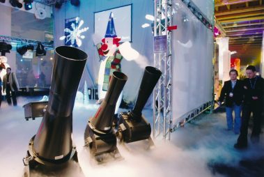 Das Bild zeigt eine Szenerie, die an eine Winterlandschaft erinnert, mit mehreren großen Maschinen im Vordergrund, die künstlichen Nebel erzeugen. Im Hintergrund steht eine bunte Figur, ein aufblasbarer Clown, der zur winterlichen oder festlichen Atmosphäre beiträgt. Die Umgebung ist mit verschiedenen Lichteffekten und Dekorationen ausgestattet. Zwei Männer gehen am rechten Bildrand entlang, was auf eine dynamische und belebte Veranstaltung hinweist. Die Mischung aus Technik und festlicher Gestaltung schafft eine eindrucksvolle Präsentation.