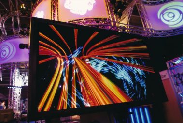 Das Bild zeigt eine beeindruckende Darstellung digitaler Kunst auf einem Bildschirm, der lebendige und dynamische Lichtstrahlen in Gelb-, Rot- und Blautönen zeigt, die sich in geschwungenen Linien bewegen. Im Hintergrund ist eine moderne Messe- oder Ausstellungsumgebung mit futuristischen Elementen wie beleuchteten Bannern und Metallgerüsten zu erkennen. Die farbenfrohen Projektionen und die geschwungene Struktur der Lichtstrahlen schaffen eine energetische und kreative Atmosphäre, die auf eine Präsentation moderner Technologie oder Kunst hinweist.
