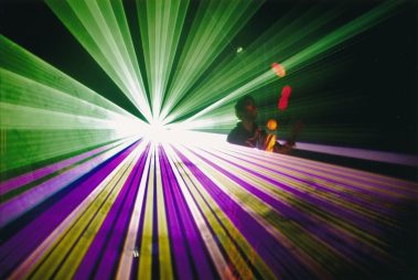Das Bild zeigt eine eindrucksvolle Lasershow, bei der grüne und violette Lichtstrahlen von einem zentralen Punkt ausstrahlen und einen intensiven visuellen Effekt erzeugen. Im Hintergrund ist die Silhouette einer Person zu sehen, die in die Show eingebunden ist, möglicherweise als Künstler oder Darsteller. Die Strahlen füllen den Raum und vermitteln ein Gefühl von Dynamik und Energie, während farbige Lichtpunkte zusätzlich die Szene bereichern. Dieses visuelle Spektakel könnte Teil einer Veranstaltung oder Präsentation sein, die auf modernste Lichttechnik setzt.