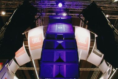 Das Bild zeigt ein beeindruckendes Lautsprecher-Array von JBL Professional, das auf einer Messe präsentiert wird. Die Lautsprecher sind vertikal gestapelt und in ein komplexes Gerüst aus Traversen eingebettet. Beleuchtungseffekte in Blau heben das Array hervor und verleihen der Szene eine futuristische Atmosphäre. Im Hintergrund sind gebogene Banner mit dem JBL-Logo und der Aufschrift „Professional“ zu sehen, die den professionellen Anspruch der Marke unterstreichen. Die Anordnung und Präsentation wirken technisch anspruchsvoll und visuell ansprechend.