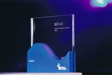 Das Bild zeigt den „Sinus – Systems Integration Award 2006“, eine elegante Trophäe, die für herausragende Leistungen im Bereich Systemintegration verliehen wird. Die Auszeichnung besteht aus einem klaren Glasblock mit eingraviertem Schriftzug, der auf einem wellenförmigen blauen Sockel ruht. Auf dem Sockel ist das Logo der Messe Frankfurt sowie die Kategorie „Business“ abgebildet. Die minimalistische Gestaltung und die hochwertige Verarbeitung betonen die Bedeutung des Preises. Der Hintergrund ist in Dunkelblau gehalten und mit sanfter Beleuchtung akzentuiert, was die Trophäe zusätzlich in den Fokus rückt und eine feierliche Atmosphäre vermittelt.