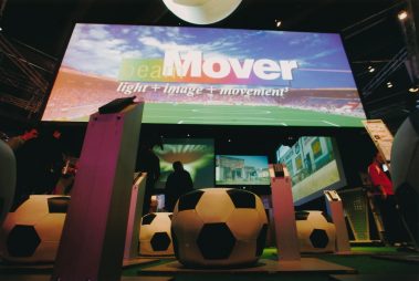 Das Bild zeigt eine Messeausstellung, die das Thema „Bewegung, Licht und Bild“ kreativ präsentiert. Im Vordergrund stehen mehrere dekorative Installationen in Form von großen Fußbällen, die auf Podesten platziert sind. Über der Szene thront ein riesiger Bildschirm, der den Schriftzug „beamMover – light + image + movement³“ anzeigt, ergänzt durch eine Stadionkulisse, die eine Verbindung zu Sport und Unterhaltung herstellt. Die Beleuchtung und die bunten Projektionen verleihen der Umgebung eine dynamische und interaktive Atmosphäre. Im Hintergrund sind weitere Displays und Messebesucher zu erkennen, die die Exponate betrachten.