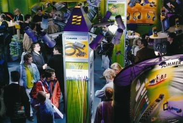 Das Bild zeigt einen Messestand des Unternehmens „Sommer Cable“ auf einer Fachmesse. Besucher bewegen sich um den Stand, betrachten ausgestellte Produkte oder führen Gespräche. Der Stand ist farbenfroh gestaltet, mit auffälligen Displays, die Kabel, Stecker und andere technische Komponenten präsentieren. Mehrere Spotlights beleuchten den Bereich, was die ausgestellten Produkte hervorhebt. Die Atmosphäre wirkt geschäftig und interaktiv, mit Besuchern, die sich über die gezeigten Lösungen informieren. Die Präsentation legt den Fokus auf die Vielfalt und Qualität von Verkabelungslösungen für professionelle Anwendungen.