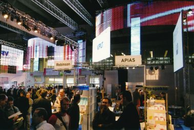 Das Bild zeigt eine lebhafte Szene auf einer Messe für Veranstaltungstechnik. Im Vordergrund sind verschiedene Messestände zu sehen, die mit Schildern wie „Robinson“ und „APIAS“ beschriftet sind. Besucher bewegen sich zwischen den Ständen, einige führen Gespräche, andere betrachten Prospekte oder informieren sich. Im Hintergrund dominiert eine große, bunte LED-Wand, die eine dynamische Atmosphäre schafft. Über den Ständen sind Traversen mit Beleuchtung angebracht, die das professionelle Messe-Setup unterstreichen. Die Szenerie vermittelt eine geschäftige, interaktive Atmosphäre, typisch für eine Fachmesse, bei der Netzwerken und Präsentationen im Mittelpunkt stehen.