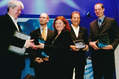 Das Bild zeigt eine Gruppe von fünf Personen bei einer Preisverleihung im Rahmen der Prolight + Sound. Eine Frau und drei Männer halten Auszeichnungen mit dem „Sinus“-Logo, die für besondere Leistungen im Bereich Systems Integration verliehen werden. Links im Bild überreicht ein weiterer Mann eine Auszeichnung, während er freundlich lächelt. Alle Anwesenden tragen formelle Kleidung, passend zum festlichen Anlass. Der Hintergrund ist in Blau und mit dem Schriftzug „Sinus“ gestaltet, was auf die Bedeutung des Moments und die professionelle Atmosphäre hinweist. Die Personen wirken stolz und erfreut über die Ehrung.