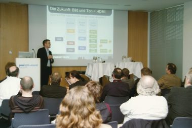 Das Bild zeigt einen Vortrag auf der Prolight + Sound, bei dem ein Referent vor einem Publikum über das Thema „Die Zukunft: Bild und Ton = HDMI“ spricht. Im Hintergrund ist eine Präsentationsfolie zu sehen, die verschiedene HDMI-Formate und technische Spezifikationen wie Auflösung, Bildfrequenz und Farbtiefe erläutert. Der Referent steht an einem Rednerpult, das mit dem Logo der Messe gekennzeichnet ist. Das Publikum sitzt aufmerksam in Reihen, während an den Seiten Stehtische mit Getränken und Mikrofonen für Diskussionen bereitstehen. Die Atmosphäre wirkt professionell und auf Wissensvermittlung ausgerichtet.