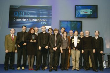 Das Bild zeigt eine Gruppe von 13 Personen auf einer Bühne, die an einer Preisverleihung teilnimmt. Im Hintergrund ist ein großes Banner mit der Aufschrift „Opus – Deutscher Bühnenpreis 2008“ zu sehen, das den Anlass verdeutlicht. Die Personen posieren gemeinsam für ein Foto, wobei eine Frau in der Mitte eine Trophäe hält, was darauf hindeutet, dass sie oder ihr Team den Preis gewonnen haben. Die Anwesenden sind überwiegend formell gekleidet, passend zur feierlichen Atmosphäre der Veranstaltung. Im Hintergrund sind außerdem mehrere Bildschirme zu sehen, die ergänzende Informationen oder Logos anzeigen. Die Stimmung wirkt professionell und freudig.