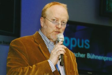 Das Bild zeigt einen Mann mittleren Alters, der in ein Mikrofon spricht, während einer Präsentation oder Moderation. Er trägt ein braunes Cord-Sakko, ein kariertes Hemd und eine Brille. Im Hintergrund ist unscharf ein Bildschirm zu erkennen, auf dem das Wort „Opus“ und „Deutscher Bühnenpreis“ zu lesen sind. Der Mann wirkt konzentriert und in die Moderation vertieft.