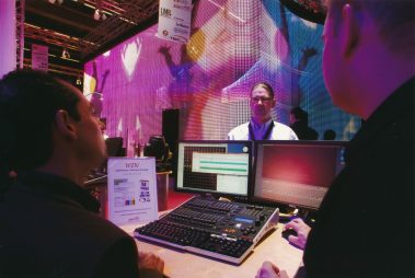 Das Bild zeigt eine Szene auf einer Messe, bei der Licht- und Bühnentechnik präsentiert wird. Im Vordergrund arbeiten zwei Personen an einem Mischpult mit Monitoren, die Lichtsteuerungssoftware anzeigen. Im Hintergrund steht eine große, farbenfrohe LED-Wand, die Animationen zeigt, und eine weitere Person im Gespräch. Die Umgebung ist in leuchtenden Farben gehalten, was die technischen Möglichkeiten der ausgestellten Produkte betont. Banner und ein Gewinnspielhinweis vervollständigen die professionelle Präsentationsatmosphäre.