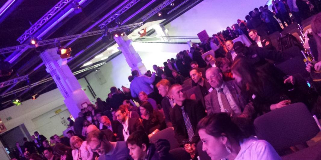 Impressionen von der Prolight + Sound 2012 Das Bild zeigt eine große Menschenmenge in einer Veranstaltungshalle. Die Sitzreihen sind gut gefüllt mit Besuchern, die sich unterhalten oder gespannt warten. Im Hintergrund stehen weitere Gruppen von Menschen, teils stehend, teils in Gespräche vertieft. Die Beleuchtung ist in warmen, lila und bläulichen Farbtönen gehalten, die dem Raum eine stimmungsvolle Atmosphäre verleihen. Die Decke ist mit Traversen und Lichttechnik ausgestattet, was auf eine professionell organisierte Veranstaltung, wie eine Messe, Konferenz oder ein Networking-Event, hinweist. Die Anwesenden sind überwiegend formell gekleidet, was die Seriosität der Veranstaltung unterstreicht.