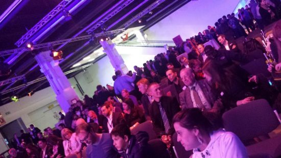 Das Bild zeigt eine große Menschenmenge in einer Veranstaltungshalle. Die Sitzreihen sind gut gefüllt mit Besuchern, die sich unterhalten oder gespannt warten. Im Hintergrund stehen weitere Gruppen von Menschen, teils stehend, teils in Gespräche vertieft. Die Beleuchtung ist in warmen, lila und bläulichen Farbtönen gehalten, die dem Raum eine stimmungsvolle Atmosphäre verleihen. Die Decke ist mit Traversen und Lichttechnik ausgestattet, was auf eine professionell organisierte Veranstaltung, wie eine Messe, Konferenz oder ein Networking-Event, hinweist. Die Anwesenden sind überwiegend formell gekleidet, was die Seriosität der Veranstaltung unterstreicht.