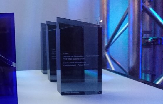 Auf dem Bild sind mehrere elegante, transparente Trophäen zu sehen, die aus Glas oder Acryl gefertigt sind. Sie stehen aufgereiht auf einer beleuchteten Fläche, was ihre klare, hochwertige Optik betont. Der Text auf den Trophäen ist teilweise lesbar und zeigt, dass sie für herausragende Leistungen in der Veranstaltungsbranche vergeben wurden. Im Hintergrund sind Metallgerüste und blaue Lichtakzente zu erkennen, die eine moderne und technisch inspirierte Atmosphäre schaffen. Dies deutet darauf hin, dass die Aufnahme im Rahmen einer Preisverleihung oder Fachmesse entstanden ist.