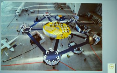 Das Bild zeigt eine beeindruckende Installation auf einem Flughafenvorfeld. Zentral ist eine gelb-blaue, kreisförmige Struktur mit mehreren verbundenen Plattformen, die durch Stege miteinander verbunden sind. Jede Plattform scheint einen bestimmten Bereich oder eine Ausstellung zu beherbergen, möglicherweise mit Sitzgelegenheiten oder interaktiven Elementen. Im Hintergrund sind Flugzeuge und Flughafenanlagen zu sehen, was den Standort eindeutig als Flughafen identifiziert. Das Design wirkt futuristisch und gut durchdacht, mit klaren Linien und einer Farbgestaltung, die an die Corporate Identity einer Fluggesellschaft erinnert. Die Installation scheint für eine Veranstaltung, Präsentation oder Messe konzipiert zu sein, um Besucher anzuziehen und eine interaktive Erfahrung zu bieten.