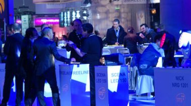 Das Bild zeigt eine lebhafte Networking-Szene auf einer Messe. Im Vordergrund stehen mehrere Personen, die in Gespräche vertieft sind, vermutlich Geschäftskontakte knüpfen oder Fachgespräche führen. Die Umgebung ist in ein stimmungsvolles blaues Licht getaucht, was eine professionelle und zugleich entspannte Atmosphäre schafft. Im Hintergrund sitzen weitere Personen an Tischen, teilweise in Gespräche vertieft oder in Ruhe ihre Umgebung beobachtend. Auf einigen beleuchteten Podesten im Vordergrund sind Produkte wie "SCENA LED 150" oder "NICK NRG 1201" ausgestellt, was auf eine Präsentation von Licht- oder Bühnentechnik hinweist. Der Stand strahlt Modernität und Fachkompetenz aus und lädt zu Interaktionen ein.