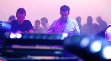 Das Bild zeigt eine dynamische Messeatmosphäre, in der zwei Männer im Vordergrund konzentriert mit Geräten oder Notizen beschäftigt sind. Die Szene ist in ein weiches, violettes Licht getaucht, was für eine kreative und technische Umgebung spricht. Im Hintergrund erkennt man schemenhaft weitere Personen, die entweder Gespräche führen oder sich die ausgestellten Exponate ansehen. Im Vordergrund sind unscharfe Lichttechnik-Geräte sichtbar, die das thematische Umfeld der Veranstaltung – vermutlich Licht- und Bühnentechnik – unterstreichen. Die Stimmung wirkt geschäftig und fokussiert, mit einem Hauch von Kreativität und Innovation.