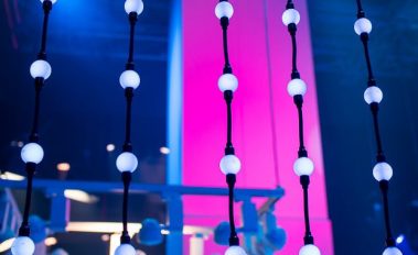 Das Bild zeigt eine künstlerische Installation aus vertikalen Lichtstreifen, die aus mehreren runden Leuchtkugeln bestehen, die an Kabeln aufgehängt sind. Die Kugeln strahlen in einem sanften Weiß und stehen im Kontrast zur dominanten Hintergrundbeleuchtung in Pink- und Blautönen. Die farbige Beleuchtung verleiht der Szene eine moderne und atmosphärische Wirkung, während die geometrische Anordnung der Leuchtelemente eine elegante Struktur schafft. Im Hintergrund sind weitere Messelemente und Ausstellungsstücke zu erkennen, was darauf hindeutet, dass die Aufnahme auf einer Veranstaltung mit Fokus auf Lichtdesign oder Bühnentechnik gemacht wurde.