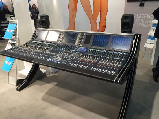 Das Bild zeigt ein hochmodernes Mischpult, das auf einem Messestand ausgestellt ist, vermutlich bei der Prolight + Sound 2016. Das Mischpult verfügt über mehrere digitale Displays, Regler und Tasten, die eine umfangreiche Steuerung ermöglichen. Es steht auf einem eleganten, geschwungenen schwarzen Standfuß, der dem Gerät ein futuristisches Design verleiht. Im Hintergrund sind ein minimalistischer Standaufbau sowie weitere Ausstellungsobjekte und Besucher zu sehen. Zwei Lautsprecher flankieren das Mischpult, was auf eine praktische Vorführung hindeutet. Die Präsentation des Geräts strahlt Professionalität aus und richtet sich an Experten aus der Veranstaltungstechnik.