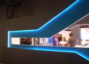 Das Bild zeigt einen modernen Messestand mit einem markanten, futuristischen Design. Die Wand des Standes ist in einem dunklen Grau gehalten und wird von einer geschwungenen, leuchtenden LED-Linie durchzogen, die in einem kräftigen Blau erstrahlt. Ein eingelassenes Fenster bietet Einblick in den Innenraum des Standes, wo mehrere Personen in ein Gespräch vertieft sind. Die ausgestellten Produkte, die möglicherweise Audiotechnik umfassen, sind im Hintergrund zu sehen. Das minimalistische und stilvolle Design des Standes zieht die Aufmerksamkeit auf sich und vermittelt einen professionellen und hochwertigen Eindruck.