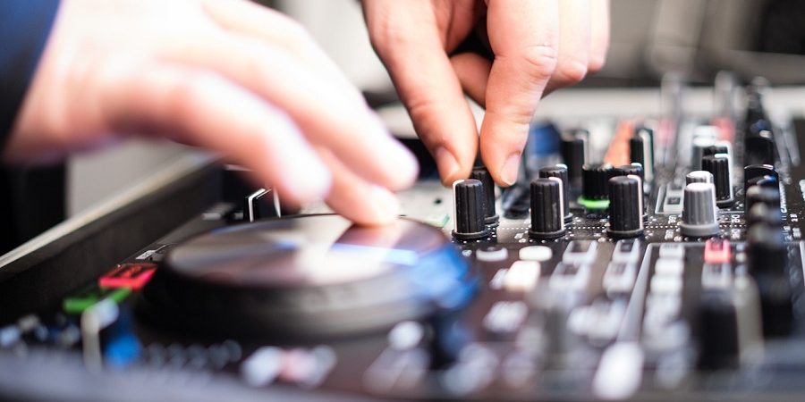 Soundcheck Mischpult Das Bild zeigt eine Nahaufnahme eines Mischpults, auf dem sich mehrere Drehregler, Schieberegler und Knöpfe befinden. Zwei Hände justieren verschiedene Einstellungen – eine Hand dreht einen Regler, während die andere anscheinend einen anderen Bereich des Mischpults bedient. Die Aufnahme ist leicht unscharf im Vordergrund, wodurch der Fokus auf die Handbewegungen und die Bedienelemente gelegt wird. Das Bild vermittelt eine dynamische Atmosphäre und suggeriert eine intensive Arbeit mit Audiotechnik, wie sie beispielsweise im MixLab auf der Prolight + Sound zum Einsatz kommt.