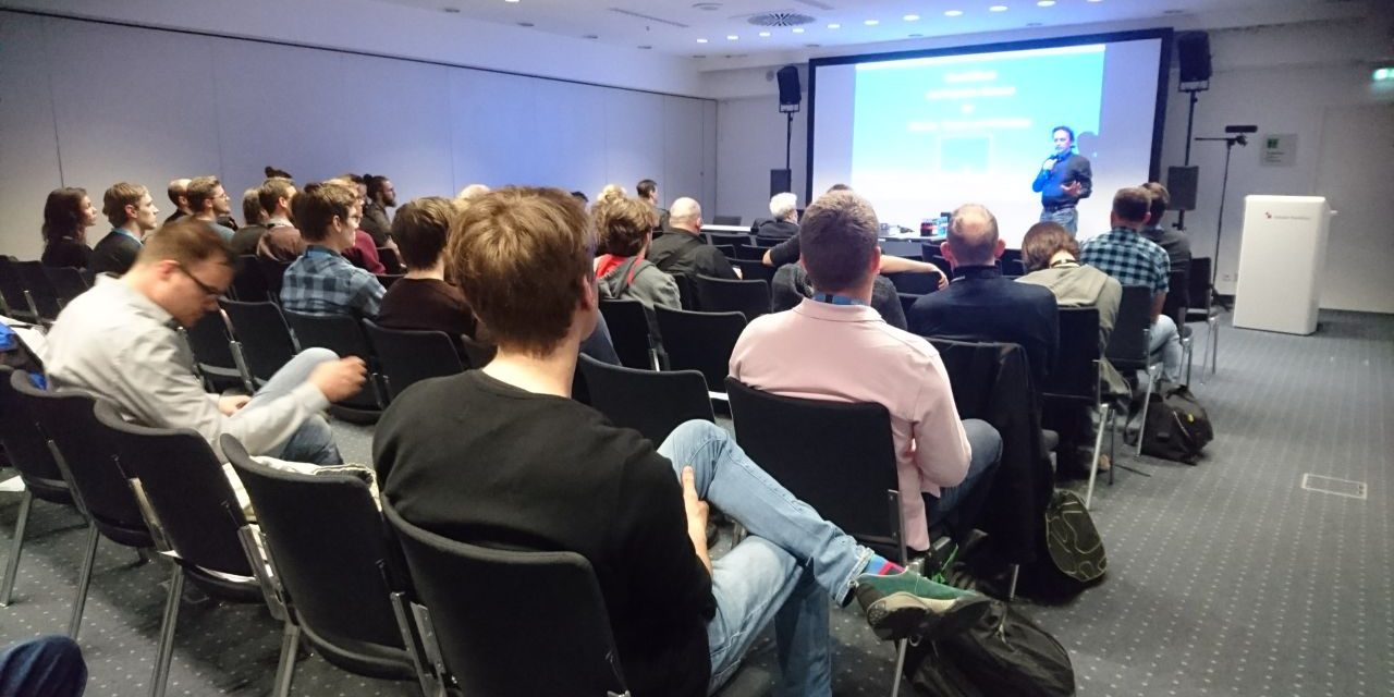 Vortrag Immersive Technologies Prolight + Sound 2018 Das Bild zeigt eine Vortragssituation in einem Konferenzraum. Die Teilnehmer sitzen auf schwarzen Stühlen in mehreren Reihen und hören einem Redner zu, der sich vorne im Raum befindet. Der Vortragende steht neben einer großen Leinwand, auf der eine Präsentation mit blauem Hintergrund und weißer Schrift zu sehen ist. Er hält ein Mikrofon in der Hand und scheint engagiert zu sprechen. Der Raum ist modern eingerichtet, mit heller Deckenbeleuchtung und weißen Wänden. Es gibt Lautsprecher an den Seiten der Leinwand und eine Kamera auf einem Stativ, was darauf hindeutet, dass der Vortrag möglicherweise aufgezeichnet oder live übertragen wird. Einige Teilnehmer tragen Namensschilder oder Konferenz-Badges, was darauf schließen lässt, dass es sich um eine Fachveranstaltung oder ein Branchenevent handelt. Die Zuhörer sind konzentriert, manche lehnen sich entspannt zurück, während andere Notizen machen oder sich mit ihren Sitznachbarn austauschen. Einige haben Taschen oder Rucksäcke neben ihren Stühlen abgestellt. Insgesamt vermittelt das Bild eine professionelle Atmosphäre und zeigt eine interessierte Zuhörerschaft in einer informativen Veranstaltung.