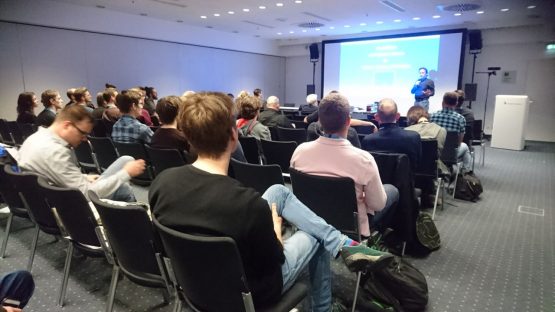 Das Bild zeigt eine Vortragssituation in einem Konferenzraum. Die Teilnehmer sitzen auf schwarzen Stühlen in mehreren Reihen und hören einem Redner zu, der sich vorne im Raum befindet. Der Vortragende steht neben einer großen Leinwand, auf der eine Präsentation mit blauem Hintergrund und weißer Schrift zu sehen ist. Er hält ein Mikrofon in der Hand und scheint engagiert zu sprechen. Der Raum ist modern eingerichtet, mit heller Deckenbeleuchtung und weißen Wänden. Es gibt Lautsprecher an den Seiten der Leinwand und eine Kamera auf einem Stativ, was darauf hindeutet, dass der Vortrag möglicherweise aufgezeichnet oder live übertragen wird. Einige Teilnehmer tragen Namensschilder oder Konferenz-Badges, was darauf schließen lässt, dass es sich um eine Fachveranstaltung oder ein Branchenevent handelt. Die Zuhörer sind konzentriert, manche lehnen sich entspannt zurück, während andere Notizen machen oder sich mit ihren Sitznachbarn austauschen. Einige haben Taschen oder Rucksäcke neben ihren Stühlen abgestellt. Insgesamt vermittelt das Bild eine professionelle Atmosphäre und zeigt eine interessierte Zuhörerschaft in einer informativen Veranstaltung.