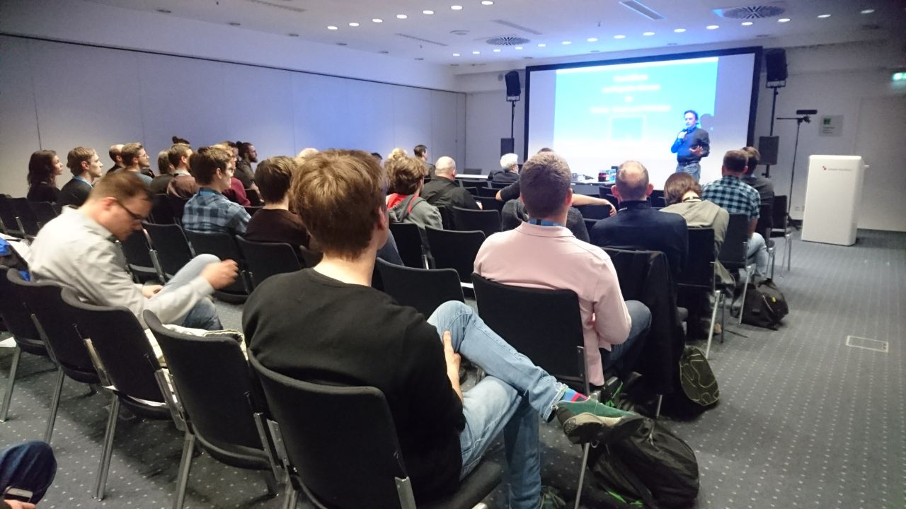 Das Bild zeigt eine Vortragssituation in einem Konferenzraum. Die Teilnehmer sitzen auf schwarzen Stühlen in mehreren Reihen und hören einem Redner zu, der sich vorne im Raum befindet. Der Vortragende steht neben einer großen Leinwand, auf der eine Präsentation mit blauem Hintergrund und weißer Schrift zu sehen ist. Er hält ein Mikrofon in der Hand und scheint engagiert zu sprechen. Der Raum ist modern eingerichtet, mit heller Deckenbeleuchtung und weißen Wänden. Es gibt Lautsprecher an den Seiten der Leinwand und eine Kamera auf einem Stativ, was darauf hindeutet, dass der Vortrag möglicherweise aufgezeichnet oder live übertragen wird. Einige Teilnehmer tragen Namensschilder oder Konferenz-Badges, was darauf schließen lässt, dass es sich um eine Fachveranstaltung oder ein Branchenevent handelt. Die Zuhörer sind konzentriert, manche lehnen sich entspannt zurück, während andere Notizen machen oder sich mit ihren Sitznachbarn austauschen. Einige haben Taschen oder Rucksäcke neben ihren Stühlen abgestellt. Insgesamt vermittelt das Bild eine professionelle Atmosphäre und zeigt eine interessierte Zuhörerschaft in einer informativen Veranstaltung.