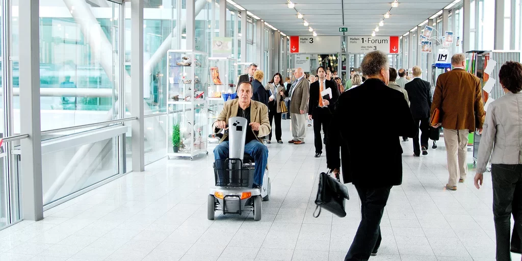 Barrierefreie Events Scooter Ein Mann fährt mit einem Elektroscooter durch einen barrierefreien Messegang mit vielen Besuchenden.