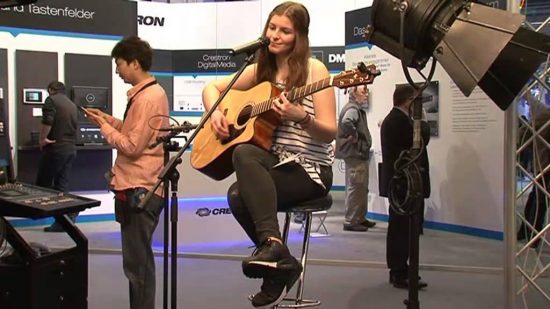 Das Bild zeigt eine junge Frau, die auf einem Barhocker sitzt und eine Akustikgitarre spielt. Sie lächelt leicht, während sie ins Mikrofon singt, das vor ihr positioniert ist. Um sie herum ist eine Messeumgebung zu sehen, mit Informationsständen und Besuchern im Hintergrund. Eine professionelle Beleuchtung, bestehend aus einem großen Scheinwerfer und weiteren technischen Geräten, hebt die Performerin hervor. Links im Bild steht ein Mann, der auf sein Handy schaut, während im Hintergrund weitere Messebesucher die ausgestellten Produkte und Displays betrachten. Die Szene vermittelt eine Mischung aus musikalischer Darbietung und technischer Präsentation, typisch für eine Veranstaltung wie die Prolight + Sound.