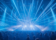 Eine große Menschenmenge steht in einer Konzerthalle und blickt auf eine Bühne, über der zahlreiche kräftige blaue und weiße Lichtstrahlen fächerförmig in den Raum schießen. Die Lichtshow erzeugt ein dichtes Netz aus Laser- und Spotstrahlen, die die gesamte Halle ausfüllen und eine dynamische, energiegeladene Atmosphäre schaffen.