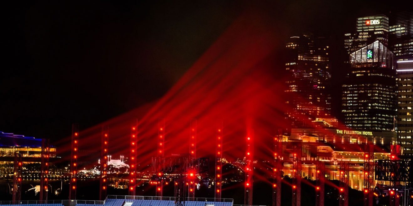 Claypaky Hy B-EYE K25 singapores-bicentennial-ndc Das Bild zeigt eine nächtliche Szene mit einer beeindruckenden Lichtinstallation. Mehrere vertikal aufgereihte rote Lichtstrahlen ragen in den Himmel und erzeugen eine dynamische Atmosphäre. Im Hintergrund sind beleuchtete Hochhäuser zu sehen, darunter das Fullerton Hotel sowie Gebäude mit markanten Logos. Die Szenerie vermittelt den Eindruck einer groß angelegten Veranstaltung, möglicherweise in einer urbanen Umgebung wie Singapur. Die Lichtinstallation dürfte von leistungsstarken LED-Moving-Heads wie dem Claypaky HY B-EYE K25 stammen, die bekannt für ihre brillante Farbintensität und Vielseitigkeit sind.
