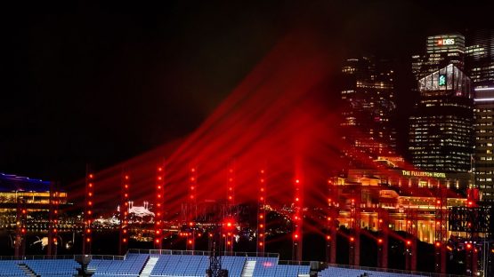 Das Bild zeigt eine nächtliche Szene mit einer beeindruckenden Lichtinstallation. Mehrere vertikal aufgereihte rote Lichtstrahlen ragen in den Himmel und erzeugen eine dynamische Atmosphäre. Im Hintergrund sind beleuchtete Hochhäuser zu sehen, darunter das Fullerton Hotel sowie Gebäude mit markanten Logos. Die Szenerie vermittelt den Eindruck einer groß angelegten Veranstaltung, möglicherweise in einer urbanen Umgebung wie Singapur. Die Lichtinstallation dürfte von leistungsstarken LED-Moving-Heads wie dem Claypaky HY B-EYE K25 stammen, die bekannt für ihre brillante Farbintensität und Vielseitigkeit sind.