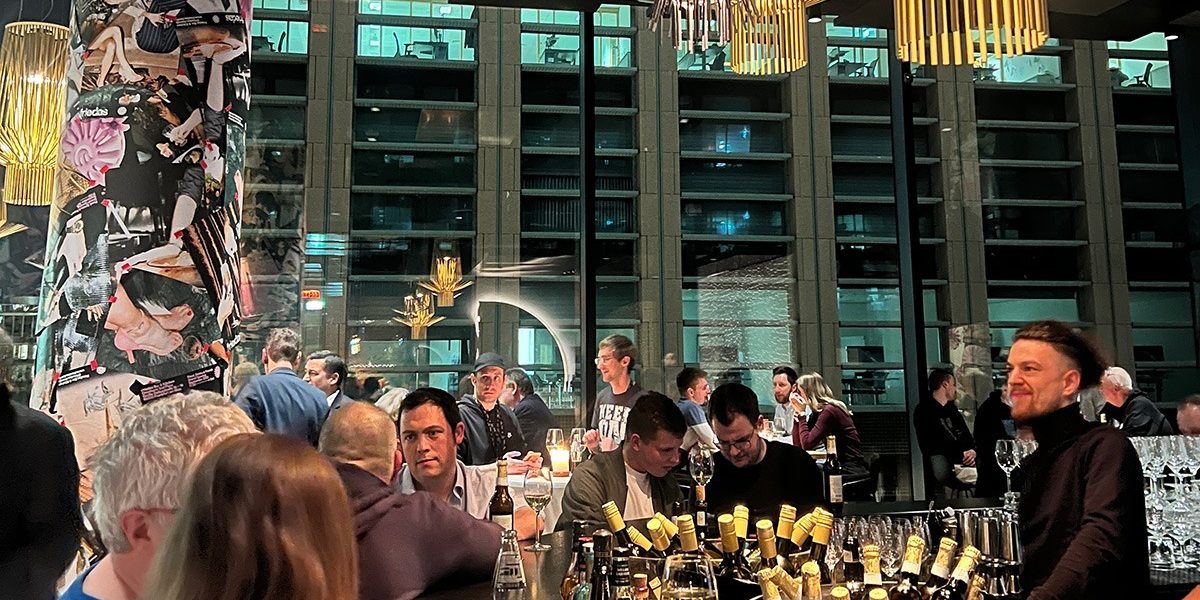 Community Nights Prolight + Sound 2024 Das Bild zeigt eine lebhafte und moderne Barszene, vermutlich von einem Networking-Event oder einer Community-Veranstaltung, bei der Menschen in entspannter Atmosphäre miteinander ins Gespräch kommen. Im Mittelpunkt steht ein großer Tresen, reich dekoriert mit Flaschen und Gläsern, während Teilnehmer in Gespräche vertieft sind oder das Ambiente genießen. Auffällige Designelemente, wie eine mit Collagen gestaltete Säule und stilvolle Beleuchtung, prägen den Raum, der durch große Fensterfronten den Blick auf ein modernes Gebäude freigibt. Die warmen Lichtakzente und die gelöste Stimmung vermitteln den Eindruck eines gelungenen Abends, der Begegnung und Austausch fördert.