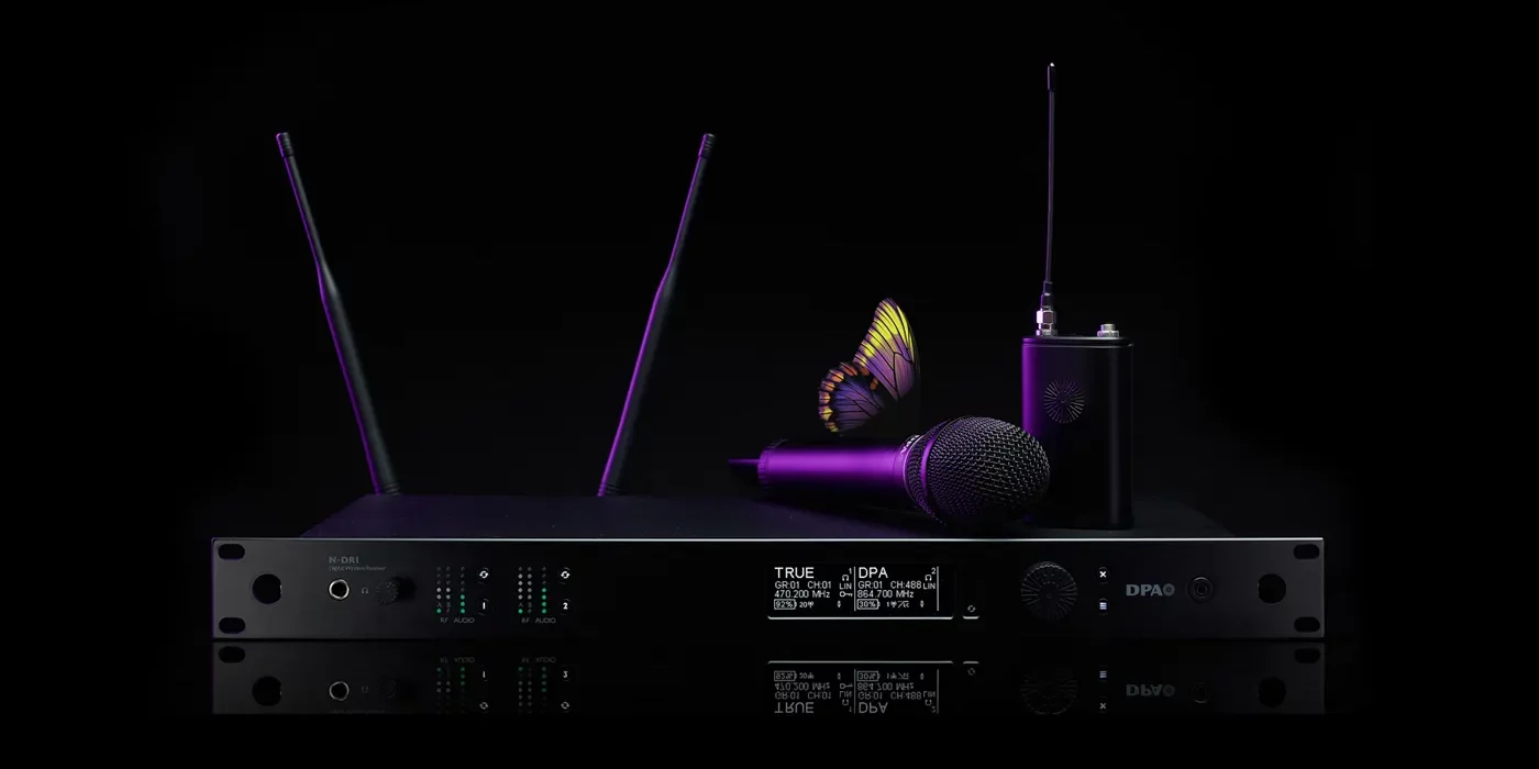 DPA N-Series Digital Wireless System Das Bild zeigt ein hochwertiges Drahtlossystem aus der DPA N-Serie Digital Wireless, bestehend aus mehreren Komponenten. Im Mittelpunkt steht ein N-DR1 Digital Wireless Receiver, ein professioneller Zweikanalempfänger mit zwei aufrecht stehenden Antennen, mehreren LED-Anzeigen und einem Display, das technische Informationen wie Frequenzen und Signalstärke darstellt. Auf dem Empfänger liegt ein kabelloses Handmikrofon, das zum DPA d:facto™-Sortiment gehören könnte. Rechts daneben steht ein Bodypack-Transmitter mit einer angeschlossenen Antenne, der eine kompakte und robuste Bauweise aufweist. Das gesamte Equipment ist in einem eleganten, schwarzen Design mit modernen, lila Akzenten gehalten. Besonders auffällig ist ein Schmetterling, der auf dem Mikrofon platziert ist. Dies könnte symbolisch für die klare und natürliche Klangqualität der DPA-Produkte stehen. Der Hintergrund ist tiefschwarz, was das Equipment in den Fokus rückt und die hochwertige Ästhetik der Produktreihe unterstreicht.