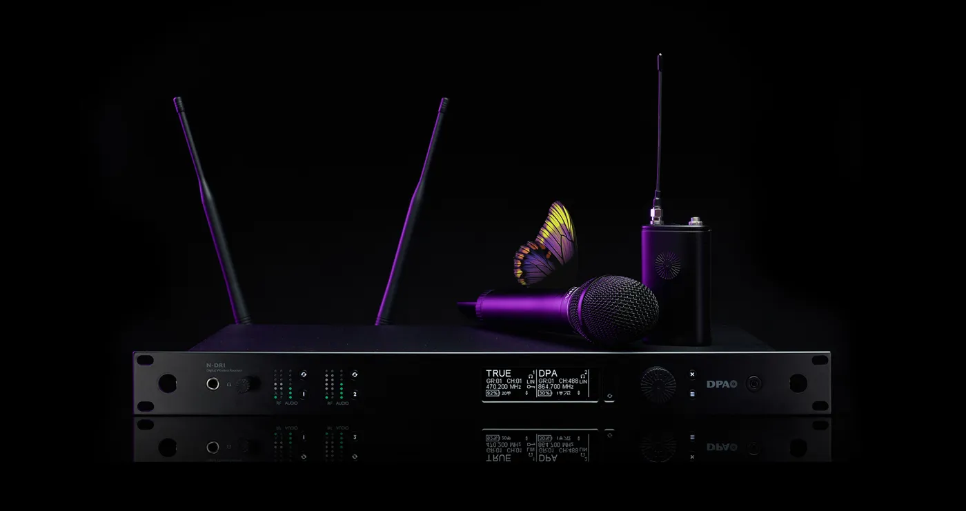 Das Bild zeigt ein hochwertiges Drahtlossystem aus der DPA N-Serie Digital Wireless, bestehend aus mehreren Komponenten. Im Mittelpunkt steht ein N-DR1 Digital Wireless Receiver, ein professioneller Zweikanalempfänger mit zwei aufrecht stehenden Antennen, mehreren LED-Anzeigen und einem Display, das technische Informationen wie Frequenzen und Signalstärke darstellt. Auf dem Empfänger liegt ein kabelloses Handmikrofon, das zum DPA d:facto™-Sortiment gehören könnte. Rechts daneben steht ein Bodypack-Transmitter mit einer angeschlossenen Antenne, der eine kompakte und robuste Bauweise aufweist. Das gesamte Equipment ist in einem eleganten, schwarzen Design mit modernen, lila Akzenten gehalten. Besonders auffällig ist ein Schmetterling, der auf dem Mikrofon platziert ist. Dies könnte symbolisch für die klare und natürliche Klangqualität der DPA-Produkte stehen. Der Hintergrund ist tiefschwarz, was das Equipment in den Fokus rückt und die hochwertige Ästhetik der Produktreihe unterstreicht.