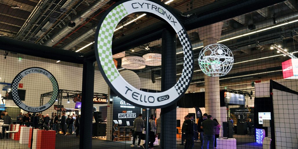 Dronen Soccer Prolight + Sound 2025 Das Bild zeigt ein Drone-Soccer-Spielfeld auf einer Messe oder Eventfläche. Im Vordergrund ist ein großer Torbogen mit der Aufschrift „CYTRONIX TELLO EDU“ zu sehen, gestaltet im typischen Schachbrettmuster. Dahinter fliegt eine Drohne in einem weißen Schutzkäfig – scheinbar auf dem Weg zum Tor oder in einer Spielszene. Um das Spielfeld herum ist ein feinmaschiges Sicherheitsnetz gespannt, das Zuschauerinnen und Zuschauer schützt. Im Hintergrund erkennt man Messestände, leuchtende Displays und Besuchergruppen, was auf eine dynamische, technikorientierte Veranstaltung hinweist. Die Szene verbindet technische Präzision mit interaktivem Messeerlebnis.