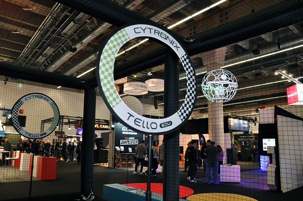 Das Bild zeigt ein Drone-Soccer-Spielfeld auf einer Messe oder Eventfläche. Im Vordergrund ist ein großer Torbogen mit der Aufschrift „CYTRONIX TELLO EDU“ zu sehen, gestaltet im typischen Schachbrettmuster. Dahinter fliegt eine Drohne in einem weißen Schutzkäfig – scheinbar auf dem Weg zum Tor oder in einer Spielszene. Um das Spielfeld herum ist ein feinmaschiges Sicherheitsnetz gespannt, das Zuschauerinnen und Zuschauer schützt. Im Hintergrund erkennt man Messestände, leuchtende Displays und Besuchergruppen, was auf eine dynamische, technikorientierte Veranstaltung hinweist. Die Szene verbindet technische Präzision mit interaktivem Messeerlebnis.