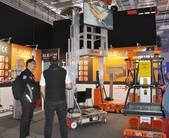 Das Bild zeigt den Messestand von Faraone auf der Prolight + Sound 2025. Im Zentrum stehen drei verschiedene Modelle der Elevah-Serie: gut sichtbar ist vorne rechts die Elevah 80 Move, rechts dahinter die Elevah 80 ES Move mit ausfahrbarem Arbeitskorb, und im linken Bildbereich die Elevah 70, ein kompakter Personenlift mit manueller Verschiebung. Zwei Besucher unterhalten sich direkt vor dem Elevah 70, während im Hintergrund ein Bildschirm eine Produktanimation zeigt. Der Stand ist in den typischen Farben Grau, Orange und Weiß gestaltet und vermittelt mit technischen Daten an den Rückwänden einen informativen Überblick über die Faraone-Produkte.