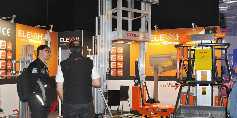 Faraone Prolight + Sound 2025 Das Bild zeigt den Messestand von Faraone auf der Prolight + Sound 2025. Im Zentrum stehen drei verschiedene Modelle der Elevah-Serie: gut sichtbar ist vorne rechts die Elevah 80 Move, rechts dahinter die Elevah 80 ES Move mit ausfahrbarem Arbeitskorb, und im linken Bildbereich die Elevah 70, ein kompakter Personenlift mit manueller Verschiebung. Zwei Besucher unterhalten sich direkt vor dem Elevah 70, während im Hintergrund ein Bildschirm eine Produktanimation zeigt. Der Stand ist in den typischen Farben Grau, Orange und Weiß gestaltet und vermittelt mit technischen Daten an den Rückwänden einen informativen Überblick über die Faraone-Produkte.