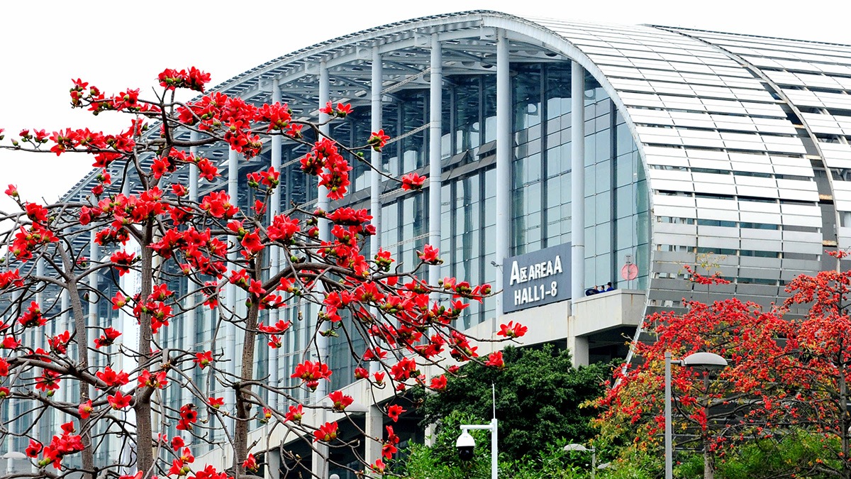 Gläserne Messehalle mit geschwungener Fassade, davor blühende rote Bäume und das Schild Area A Hall 1–8.