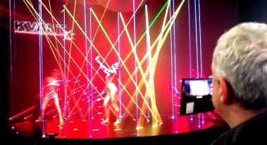 Das Bild zeigt eine dynamische Bühnenszene mit einer beeindruckenden Lichtshow. Bunte Laserstrahlen in verschiedenen Farben wie Rot, Gelb und Grün durchziehen den Raum und erzeugen eine futuristische und energiegeladene Atmosphäre. Im Hintergrund sind Tänzer oder Performer zu erkennen, die sich im Zusammenspiel mit den Lichteffekten bewegen. Im Vordergrund beobachtet ein Mann die Szenerie durch einen Bildschirm, was darauf hindeutet, dass die Performance aufgenommen oder gesteuert wird. Der Schriftzug "KVANT" deutet auf einen Hersteller oder Anbieter der Laser-Technologie hin. Die Inszenierung wirkt modern und kreativ, ideal für eine Messe oder Veranstaltung mit Fokus auf Licht- und Bühnentechnik.