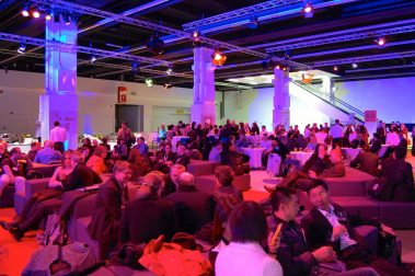 Das Bild zeigt eine lebendige Networking-Veranstaltung in einer modernen Halle mit stilvoller Beleuchtung. Verschiedene Gruppen von Personen sitzen auf gemütlichen Sitzgelegenheiten oder stehen an runden Tischen, während sie sich unterhalten und Getränke genießen. Die Atmosphäre ist entspannt, mit einem Fokus auf Austausch und Gespräche. Die farbige Beleuchtung in Blau- und Rottönen verleiht dem Raum eine warme, einladende Stimmung. Im Hintergrund sind Elemente wie hohe Säulen und eine Rolltreppe sichtbar, die die Größe der Location betonen. Die Szenerie vermittelt das Bild eines gut besuchten Events, bei dem Kontakte geknüpft und Diskussionen geführt werden.