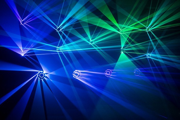 Das Bild zeigt eine beeindruckende Lichtinstallation mit mehreren Lichtquellen, die intensive blaue und grüne Strahlen in verschiedene Richtungen projizieren. Die Lichtstrahlen sind symmetrisch und überlappen sich in der Luft, wodurch ein komplexes, fast dreidimensionales Muster entsteht.Die Leuchten selbst scheinen spezielle IVL-Lichtquellen zu sein, die für immersive Lichtshows entwickelt wurden – eine Technologie, für die Minuit Une bekannt ist. Die Strahlen wirken besonders dynamisch, da sie sich in unterschiedliche Richtungen ausbreiten und den Raum vollständig einhüllen. Diese Art der Beleuchtung eignet sich hervorragend für Clubs, Live-Shows und Veranstaltungen, die eine fesselnde visuelle Atmosphäre schaffen möchten. Die Kombination aus Blau- und Grüntönen sorgt für eine futuristische und energiegeladene Ästhetik.